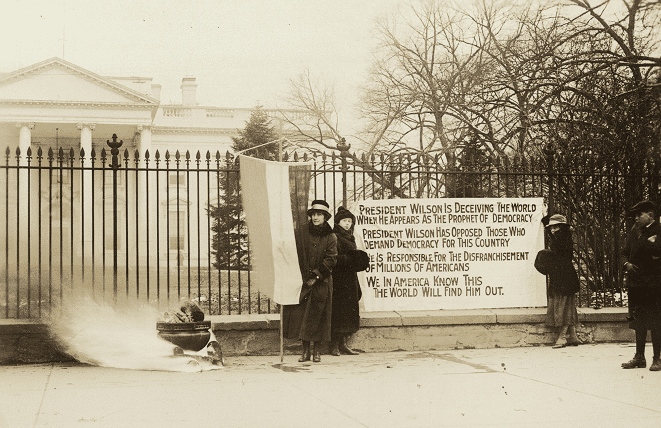 Photograph of National Woman's Party watchfire demonstrators standing with banners and fire in urn in front of White House. One banner reads, "President Wilson is deceiving the world when he appears as the prophet of democracy. President Wilson has opposed those who demand democracy for this country. He is responsible for the disfranchisement of millions of Americans. We in America know this. The world will find him out."