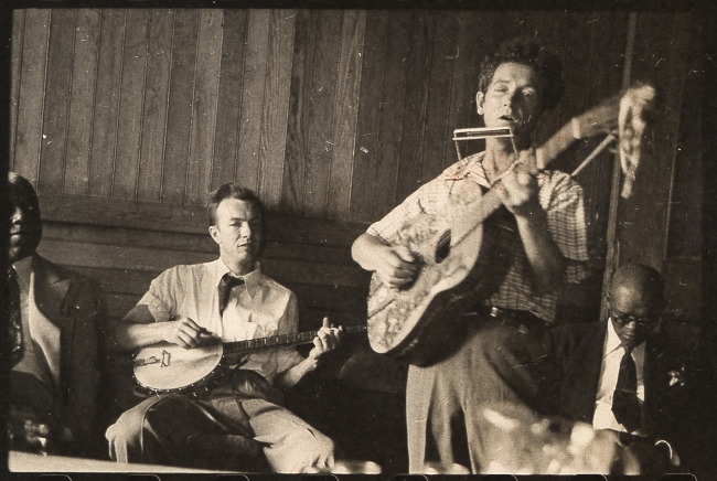 Woody Guthrie plays his guitar. Pete Seeger accompanies on banjo; music journalist Dan Burley sits at left. By Leonard Rosenberg