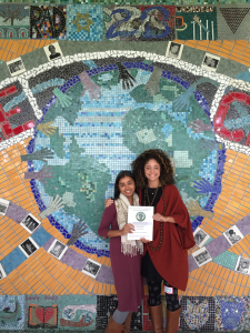 Two women smile and pose in front of a mosaic of the Earth