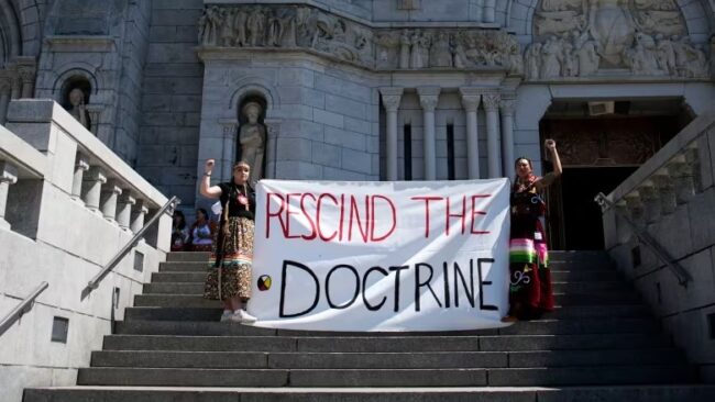 Sarain Fox, right, and Chelsea Brunelle of the Batchewana First Nation unfurled a banner reading 'Rescind the Doctrine' outside the mass presided over by Pope Francis at the National Shrine of Sainte-Anne-de-Beaupré in Quebec on July 28, 2022.